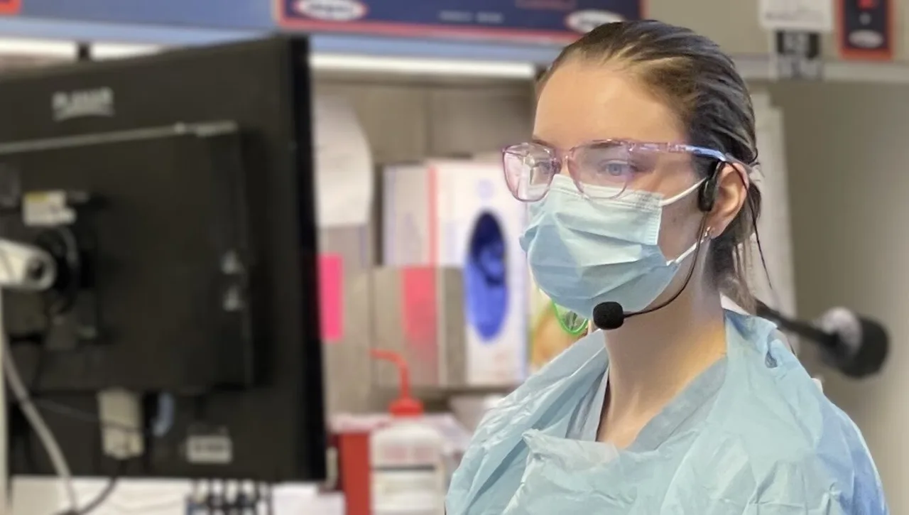 A pathologists' assistant in gowning and eye protection wearing a Sennheiser ME3 headset while dictating at a gross-room workstation